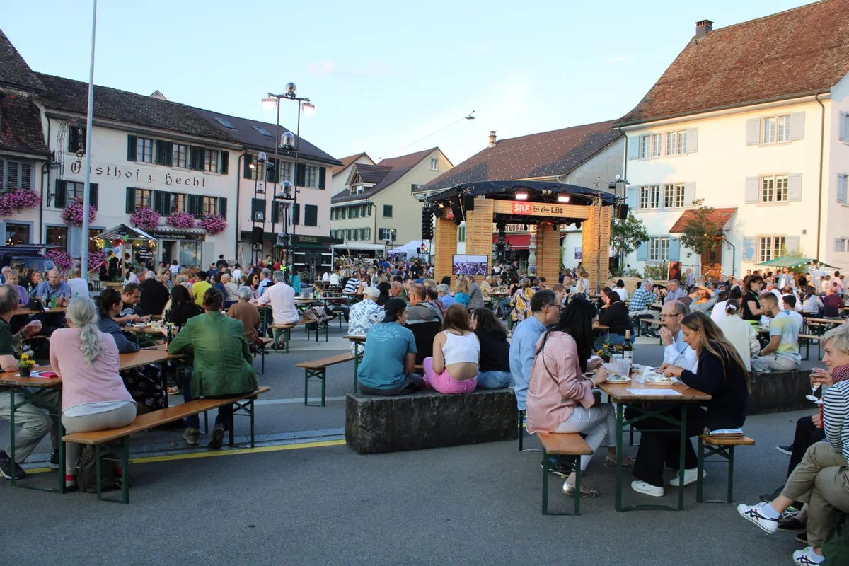 srf2_preview Viele Besucherinnen und Besucher waren schon vor Drehbeginn vor Ort., Einlass erhielt man nur mit einem Covid-Zertifikat. Es bildete sich vor dem Eingang eine Schlange., In den Anlass integriert war auch das Forellenfest des Verkehrsvereins., Dieser hatte ein eigenes Festzelt auf dem Gelände., Die Teller sind bereits vorbereitet., Die erste Forelle des Abends landet auf dem Teller., Geübt werden Gräten und Kopf entfernt..., ... entsorgt., Wer keinen Fisch mochte, durfte sich in den Restaurants oder am Grillstand verpflegen., Einlass gab es für maximal 1500 Personen., Letzte Vorkehrungen vor den Dreharbeiten.