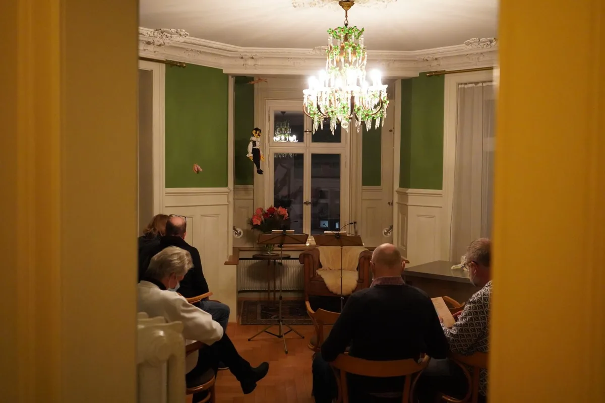 Durch eine geöffnete Tür sieht man in das Wohnzimmer der Alten Kanzlei in Wald, wo das Publikum auf Holzstühlen sitzt und auf den Beginn der Veranstaltung wartet.