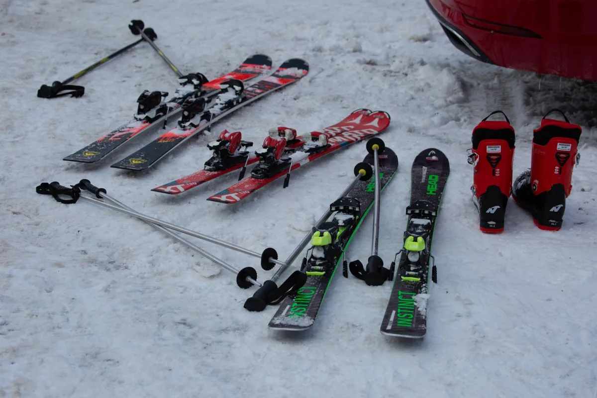 Auf dem Boden unterhalb des Kofferraums eines Wagens liegen Ski und Stöcke. Daneben steht ein paar Skischuhe