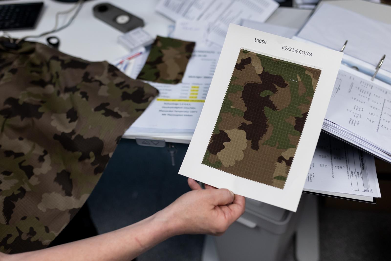 Eine Hand hält ein Blatt mit Tarnmuster hoch, im Hintergrund sieht man Papierbündel und eine Stoffprobe.