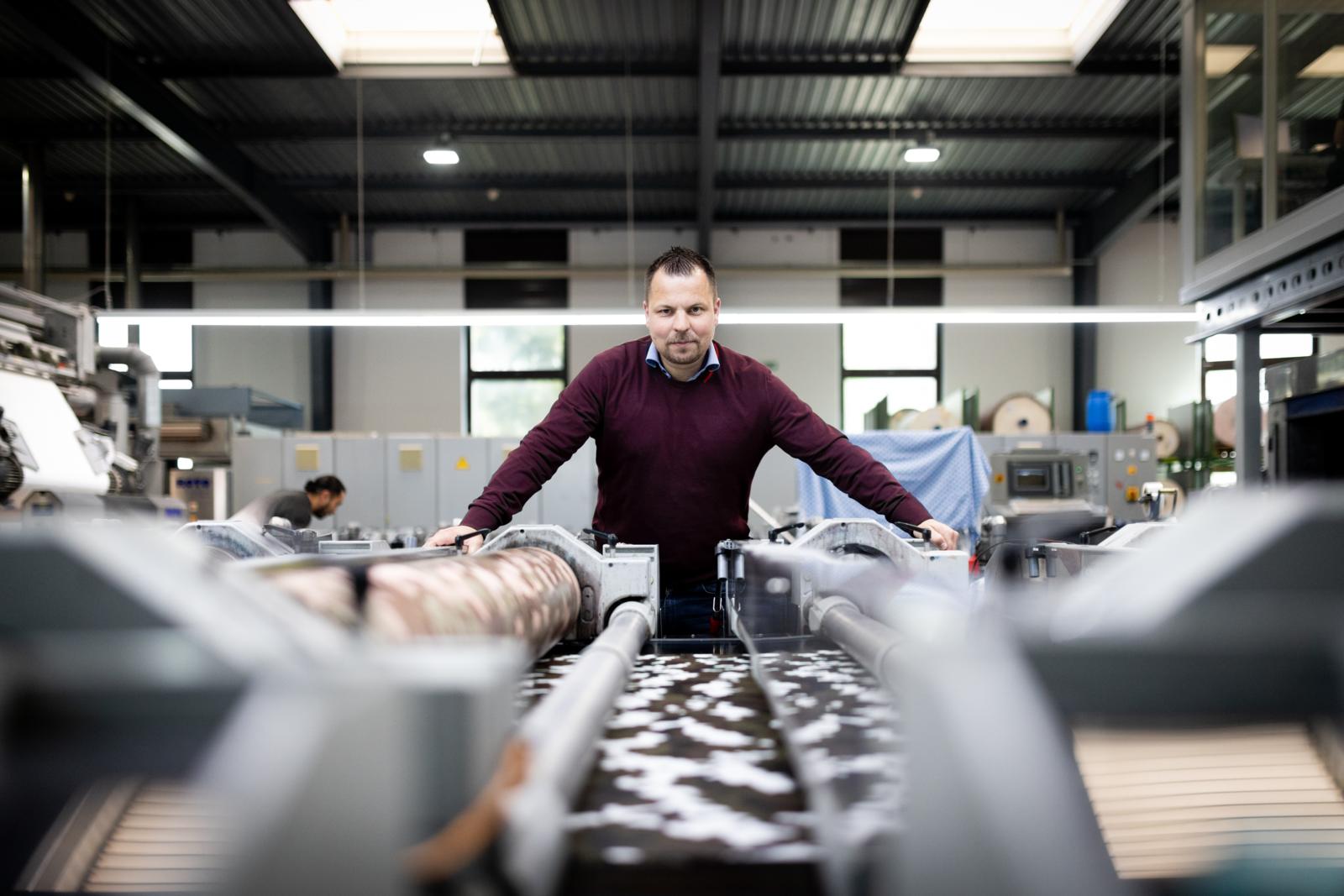 Ein Mann steht vor einem Laufband mit mehreren Walzen, durch das Armee-Stoff läuft.