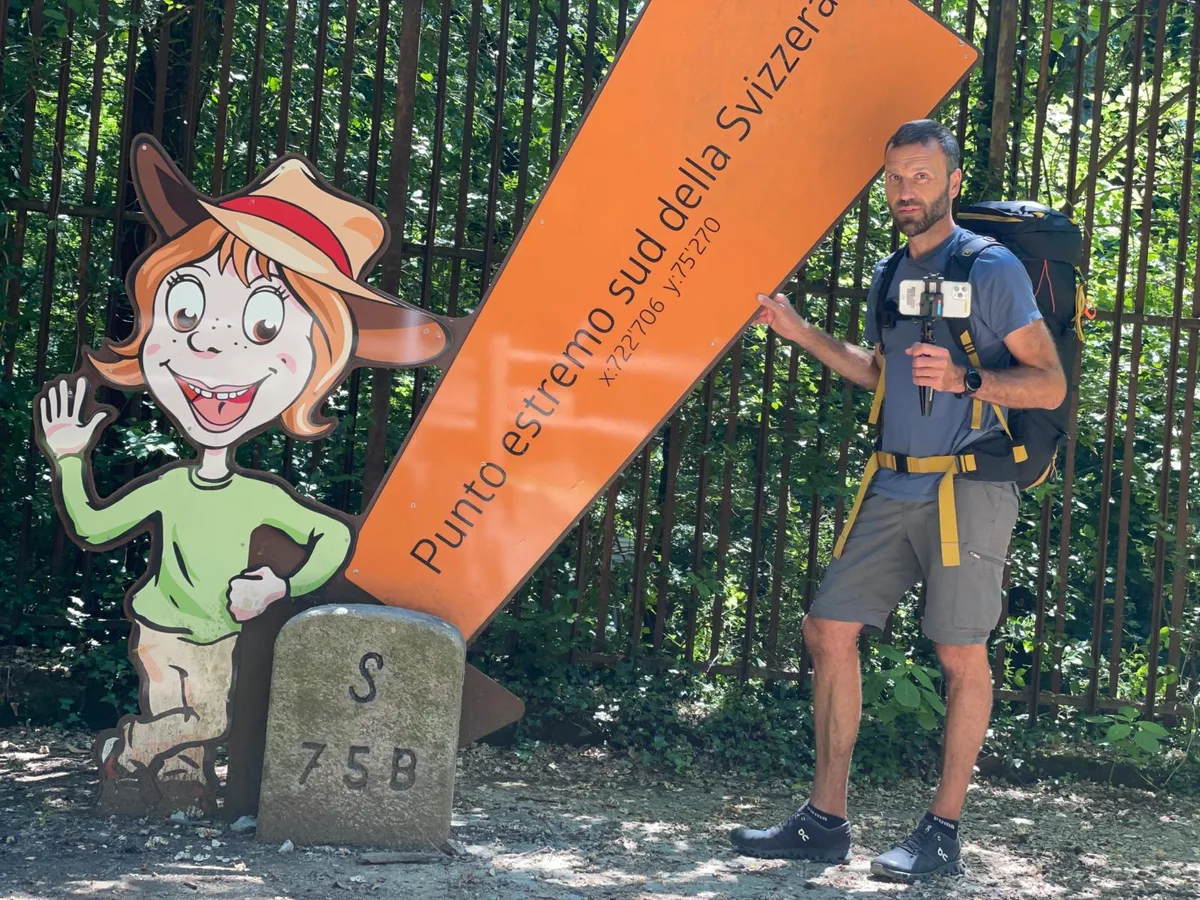 Das Ende der Reise war in Chiasso, am südlichsten Punkt der Schweiz. (Foto: PD) Mann mit Wanderausrüstung steht neben einer Holzfigur und einem Schild.