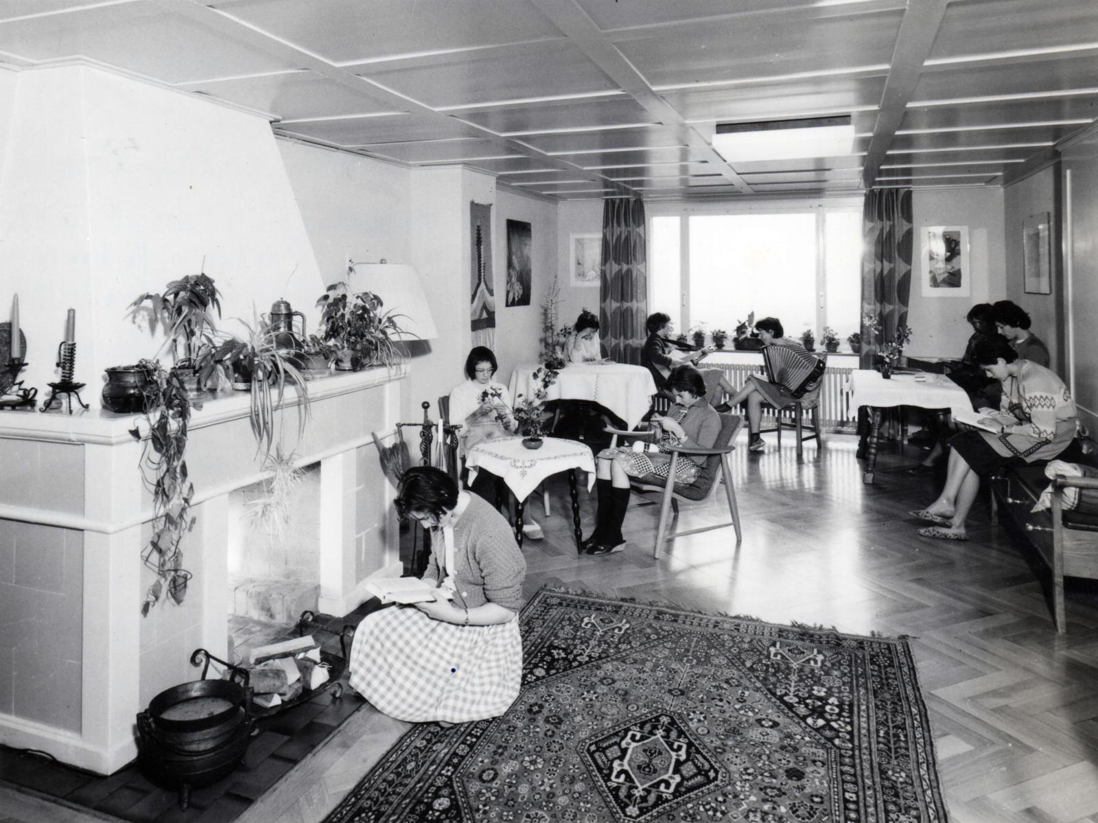 Auf dem Schwarzweiss-Foto sieht man ein grosses Wohnzimmer. Darin sitzen junge Frauen auf verschiedenen Stühlen und vor dem Kamin.