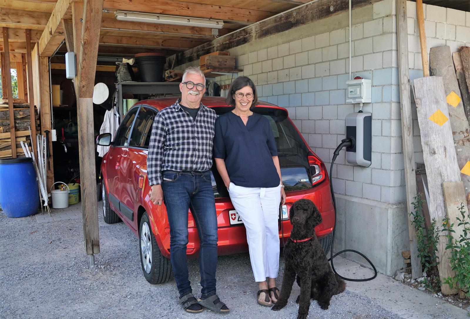Man sieht Ueli und Susanne Erb vor ihrem roten Elektroauto, das an den Strom angeschlossen ist.