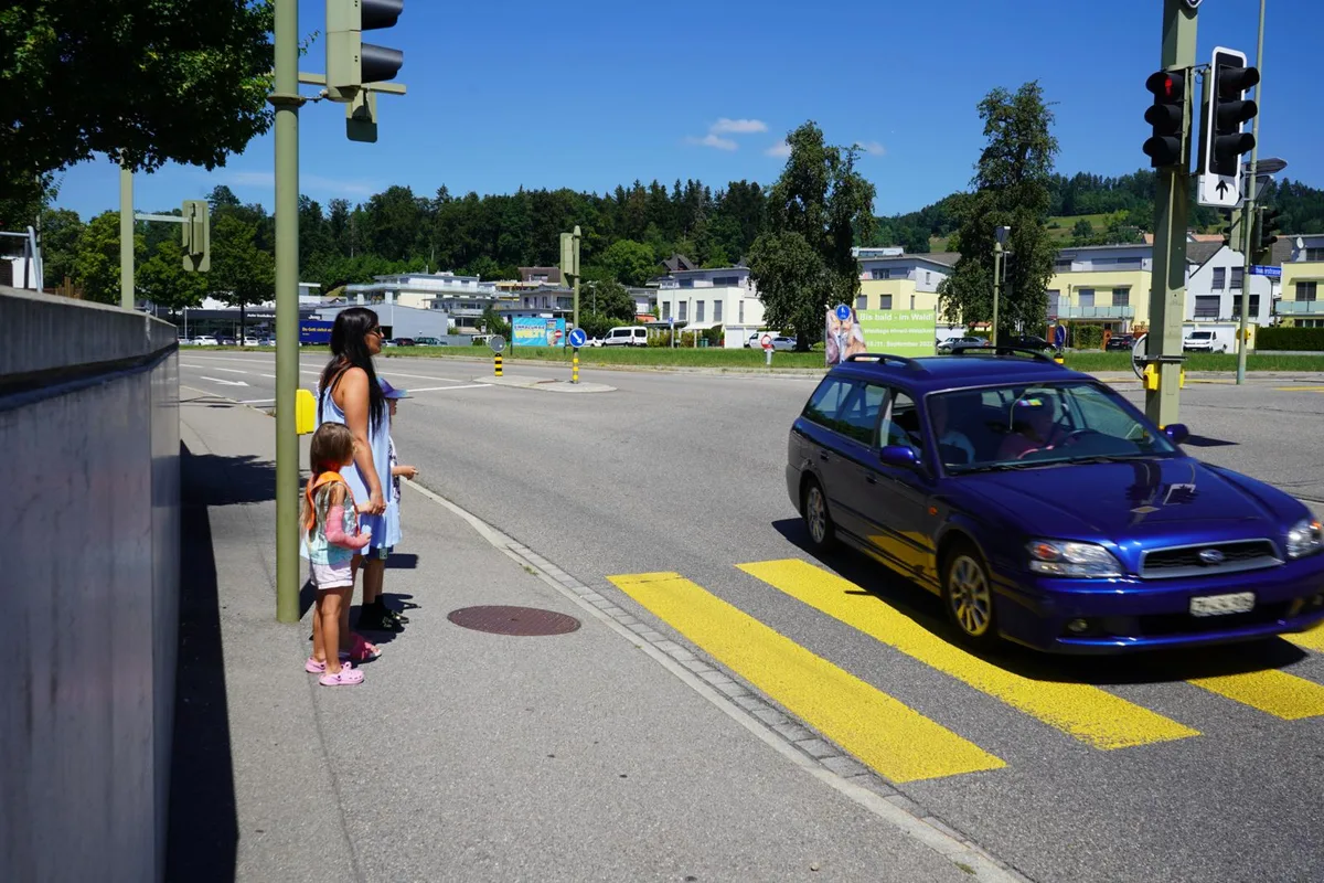 Frau steht mit zwei Kindern an einem Fussgängerstreifen während ein blaues Auto vor ihr vorbeifährt. 