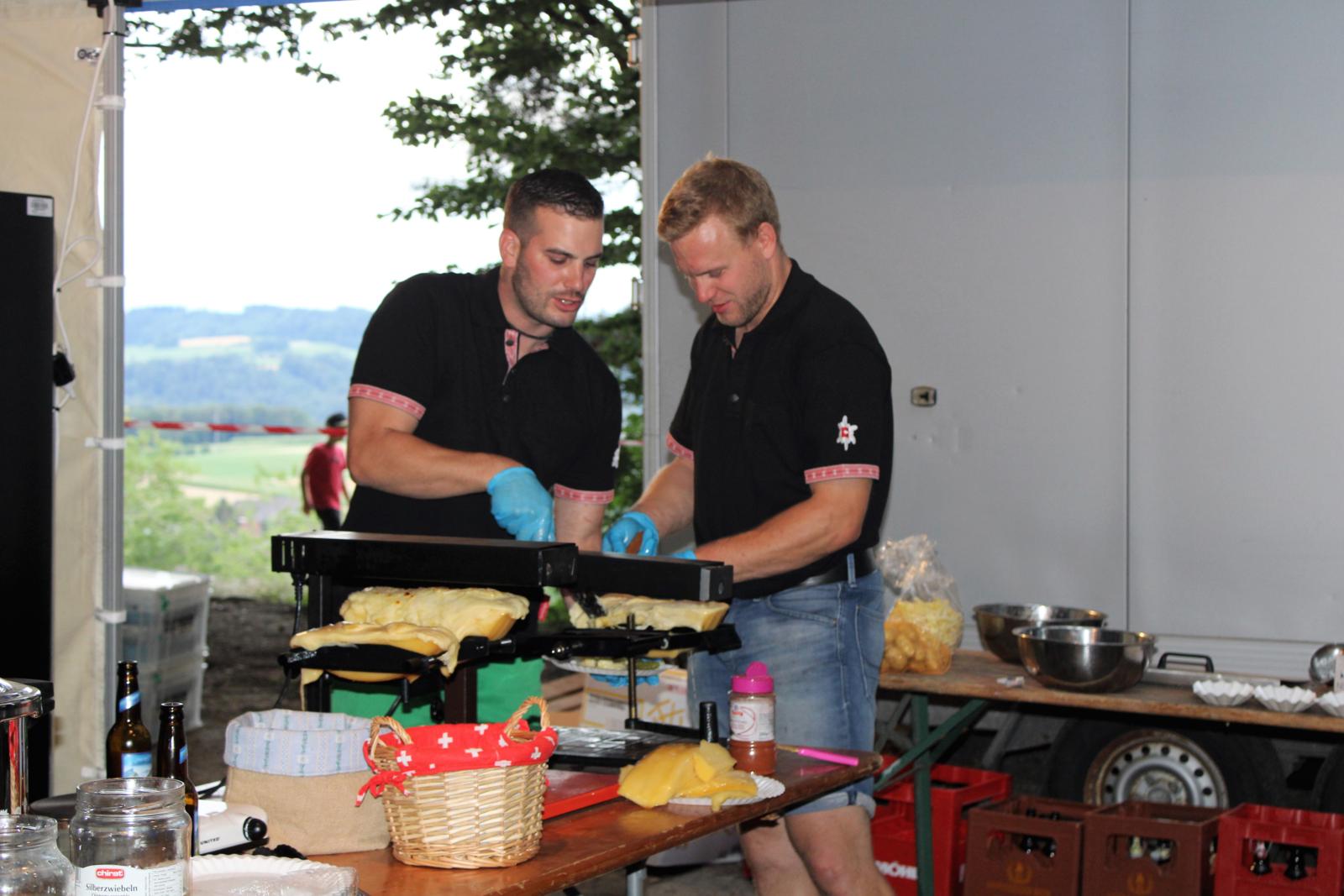 Man sieht zwei Personen, die ein Raclette vorbereiten.