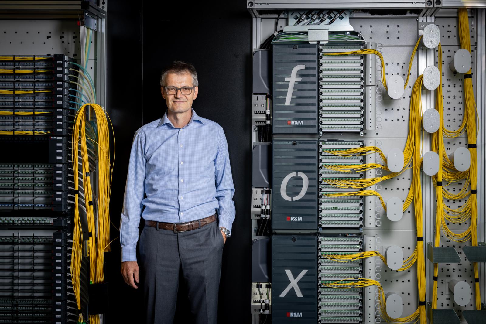 Ein Mann im blauen Hemd steht vor einem Netzwerkschrank.