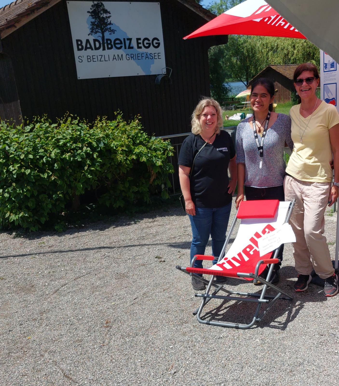 Drei Frauen stehen vor einer Holzbaracke und hinter einem Liegestuhl.