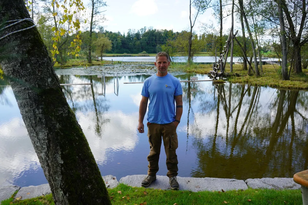 Ein Mann in blauem T-Shirt steht vor einem Weiher mit Seerosen in dem sich der Himmel spiegelt.