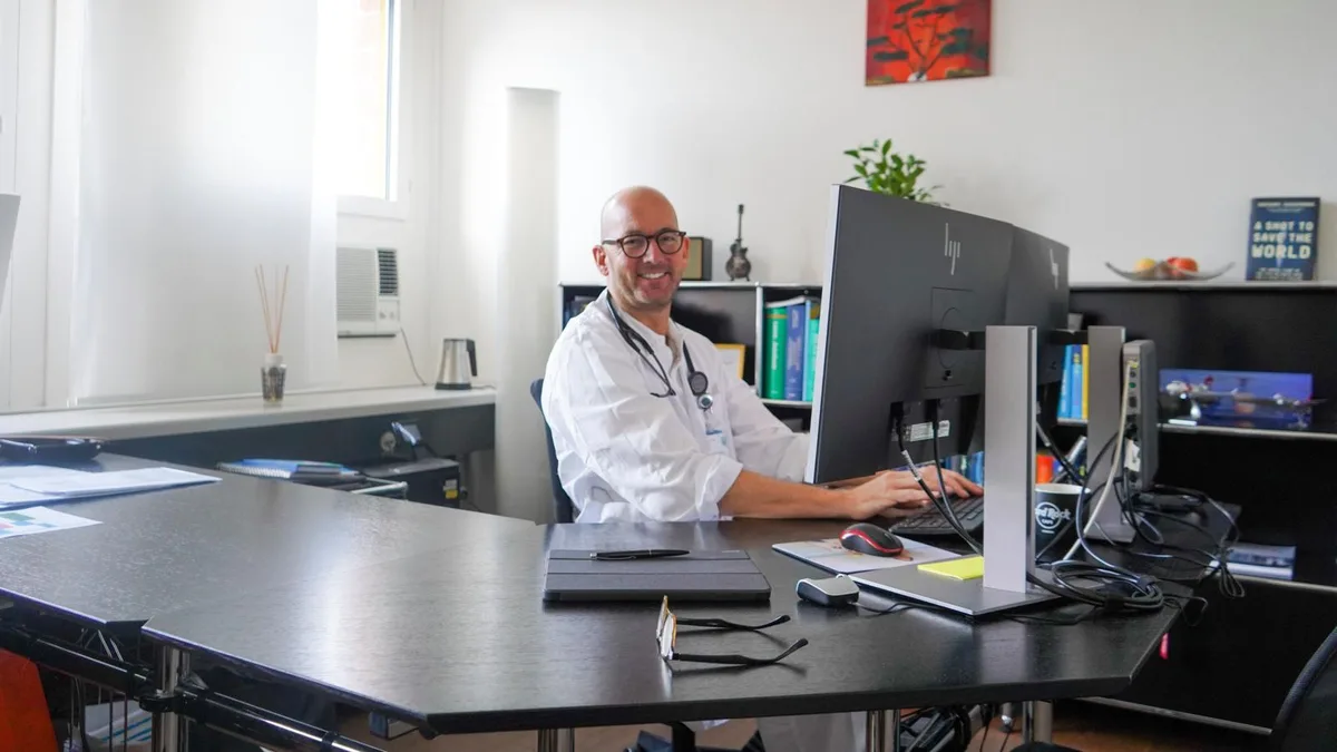 Ein Mann im weissen Kittel sitzt in seinem Büro und lacht in die Kamera. 