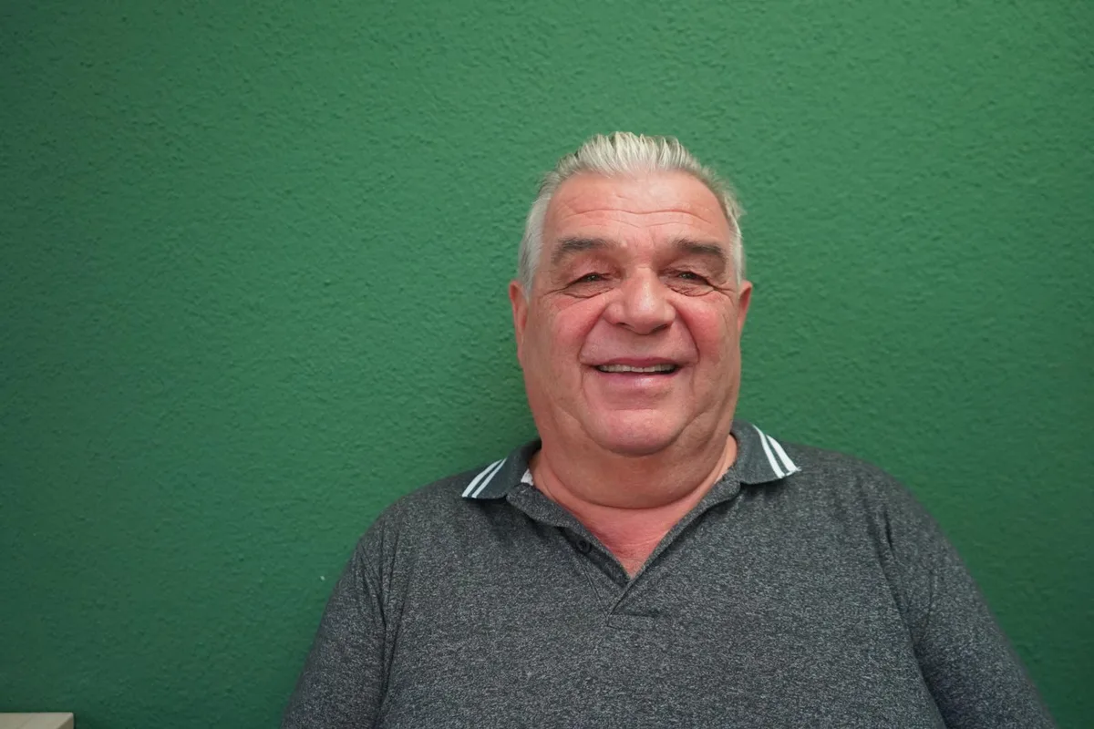 Ein Mann mit grauen Haaren sitzt vor einer grünen Wand und lacht in die Kamera. 