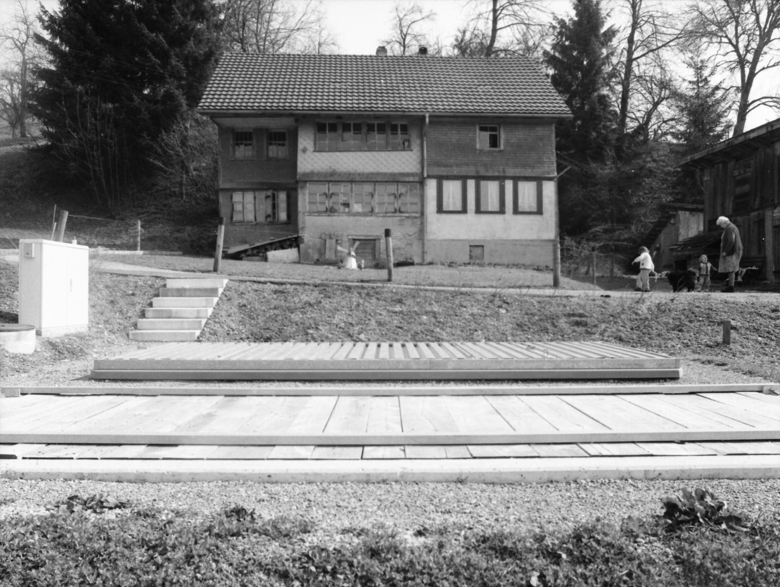 Man sieht eine Schwarz-weiss-Aufnahme von einem alten Haus, am Boden davor deutet eine Bodenplatte die Kleinkläranlage an.