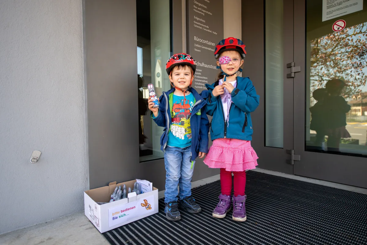 Die beiden Kinder freuen sich, dass sie das Versteck vor der Gemeindeverwaltung entdeckt haben., Ein Schokoladenhase pro Kind – mehr erlauben die Eltern nicht., Was die Eltern und die Kinder nicht wissen, hinter dem Gebäude steht ein unberührter Karton voller Hasen vor der Kellertür., 4000 Hasen waren es zu Beginn der Suche., Geplündert ist bereits der Karton vor der Kirche. , War's der Pfarrer?, Auch beim Laden der Müller und Jusell AG gibt's Osterhasen. Wo, wird nicht verraten., Die Osterhasen sind im ganzen Dorf verstreut., Das kann zu Missverständnissen führen.