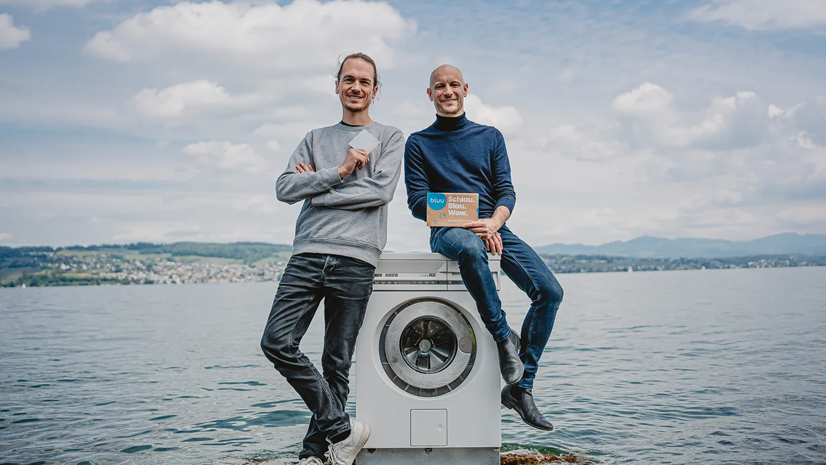 Das Gründerteam von bluu, Roman Stämpfli (links) und Marko Vidmar, steht an einem See und posiert neben einer Waschmaschine. In der Hand halten sie die bluu-Waschstreifen.