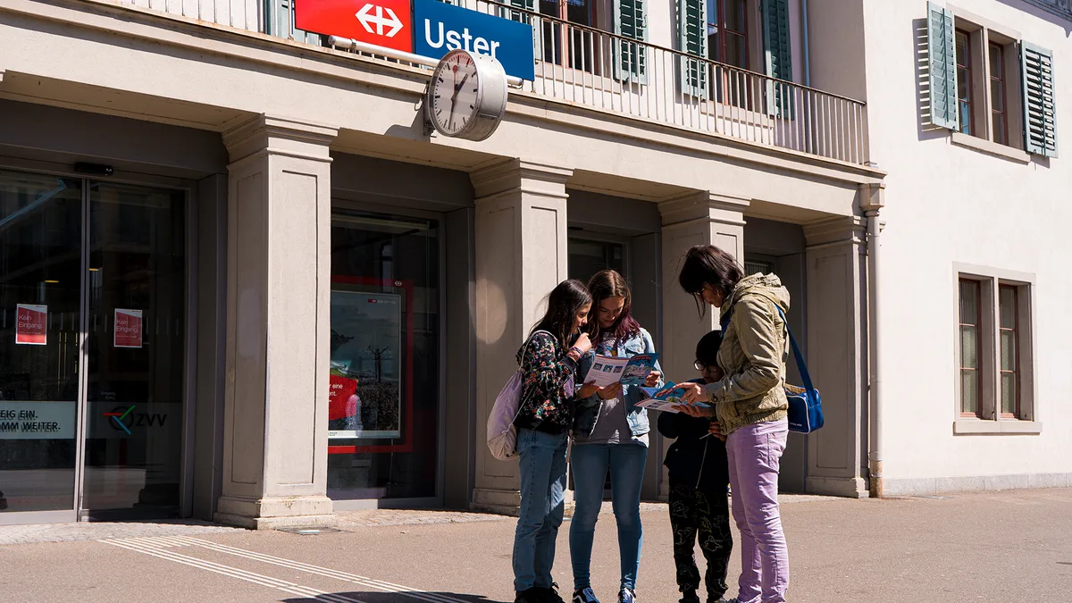 Die Reisegruppe studiert beim Bahnhof Uster die Ringo-Broschüre. Im Hintergrund ist das Bahnhofsgebäude zu sehen. 