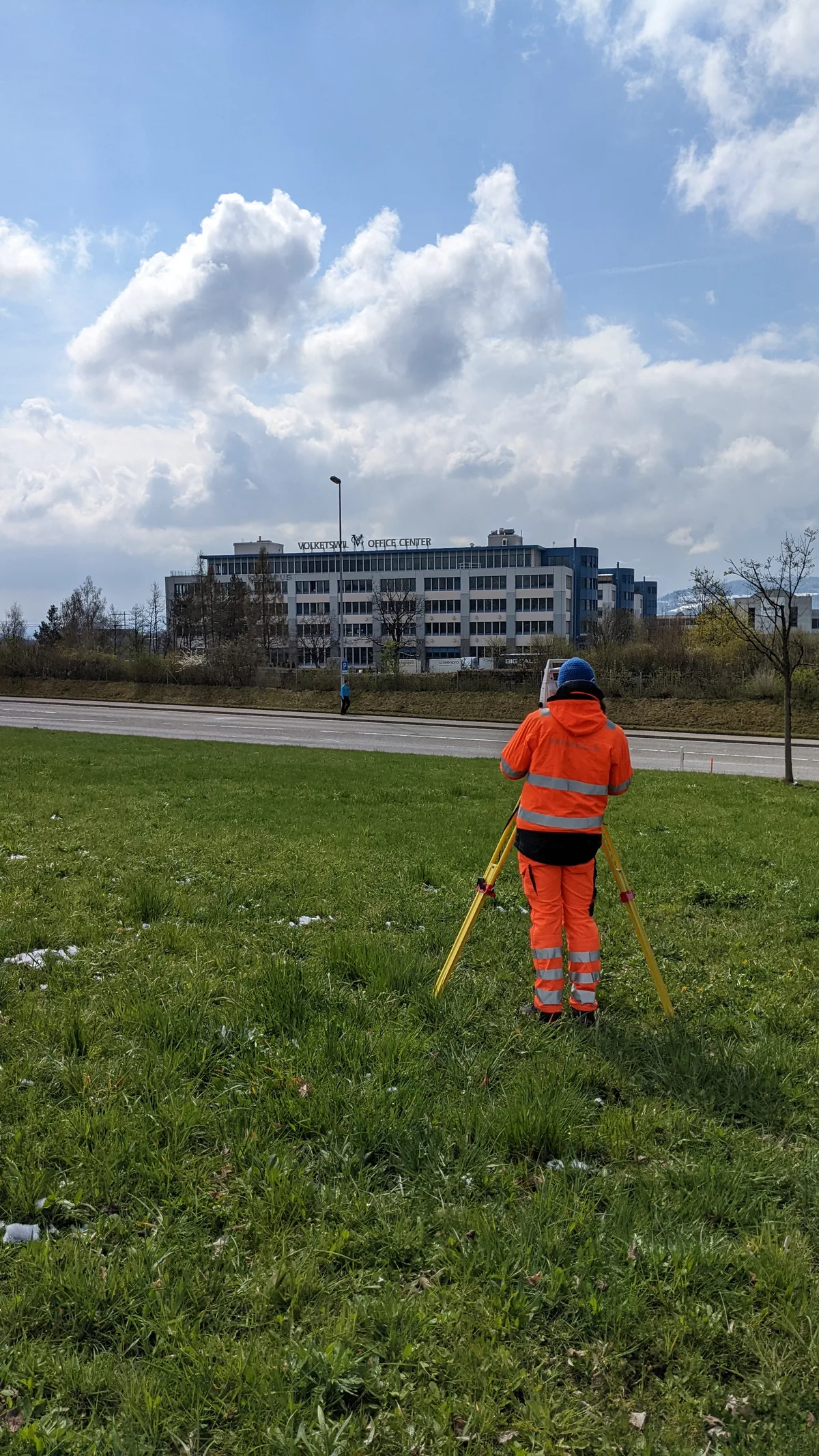 Ein Mann mit Leuchtweste steht vor einem Vermessungsgerät auf einer Wiese.