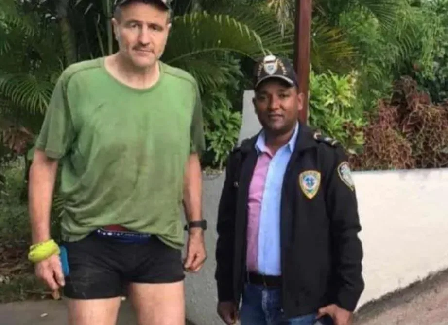 Ein Mann mit grünem T-Shirt und kurzer schwarzer Turnhose. Rechts von ihm ein etwas kleinerer Polizist.