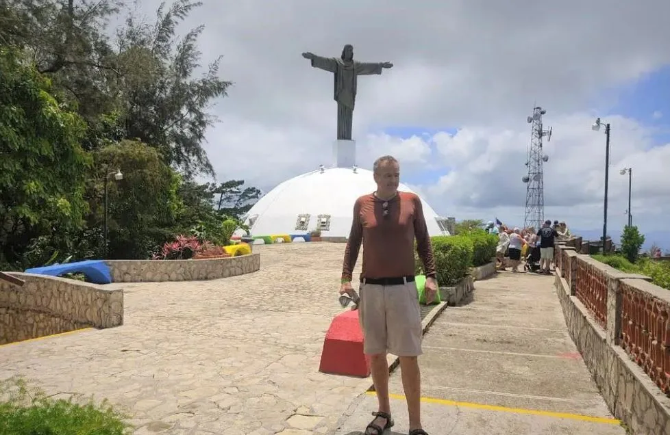Ein Mann mit braunem Shirt und kurzer Hose steht auf einer Aussichtsterrasse. Im Hintergrund eine grosse Christusstatue.