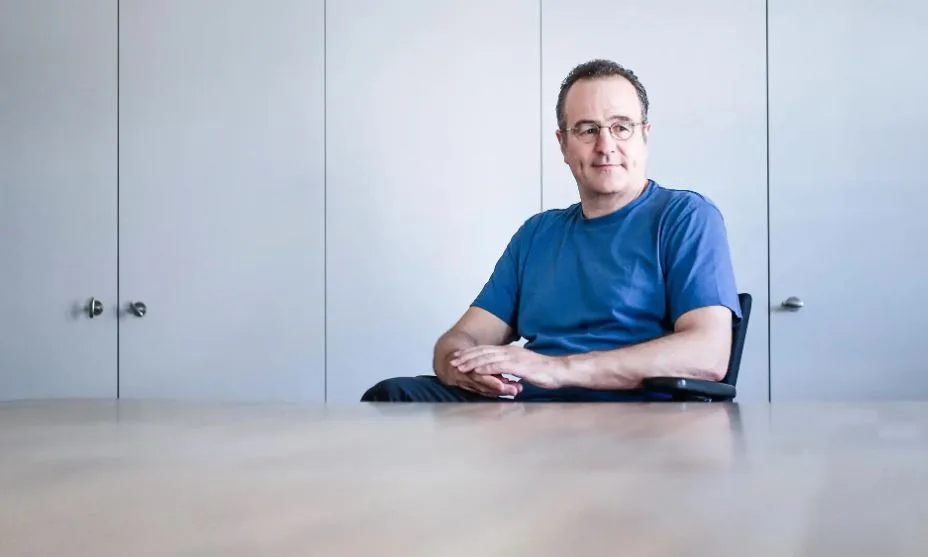 Mann mit Brille und blauem Shirt sitzt an einem Tisch und schaut direkt zum Fotografen.