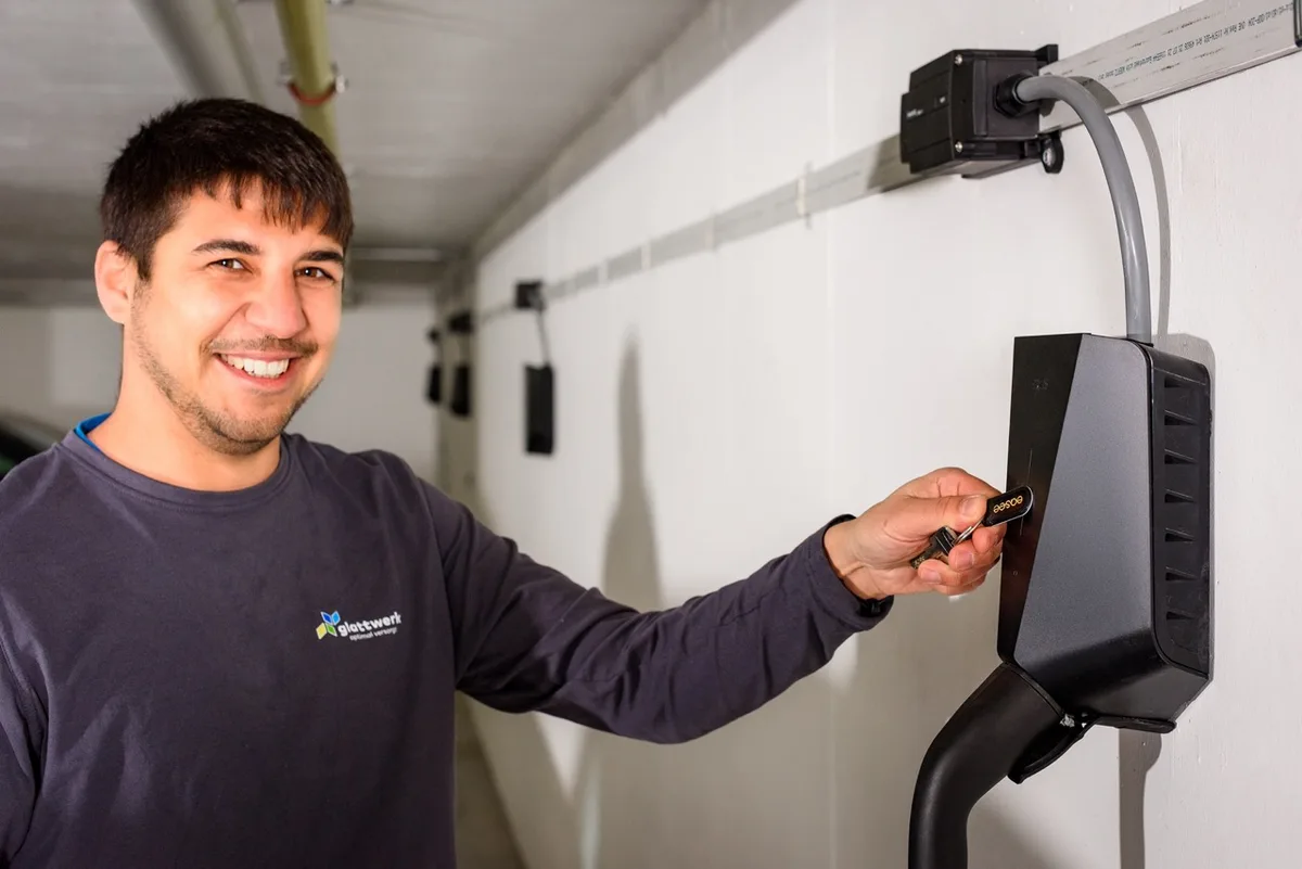 Zu sehen ist ein Installateur, der mit einer Schlüssel-ID-Karte vor einer E-Auto-Ladestation steht.
