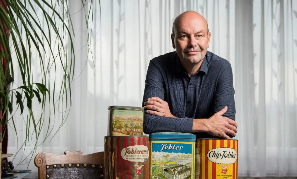 Ein Mann mit Halbglatze schaut in das Objektiv des Fotografen. Die Ellenbogen hat er auf alten Blechbüchsen abgestützt. Auf ihnen steht Tobler beziehungsweise Toblerone.