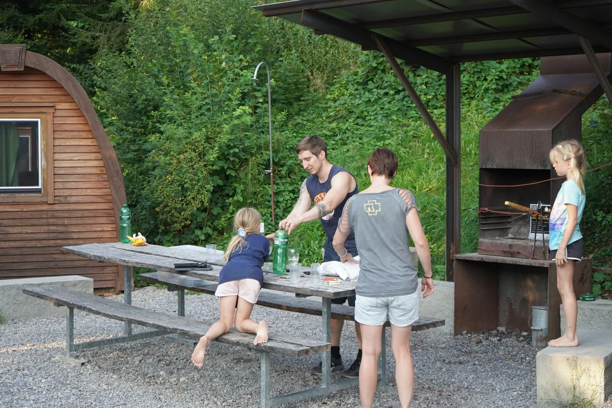 Essen zwischen Holz-Iglus: Familie Gremminger an ihrem ersten Ferienabend.., Die Familie verbringt ihre Sommerferien auf dem Campingplatz Atzmännig., Dort schlafen sie drei Nächte in einem der Podhäuser., Die Unterkünfte können gemietet werden., Auf einer kleinen Lichtung stehen mehrere der Holz-Iglus, die mit Betten, Strom, Licht und sogar einer Heizung ausgestattet sind, Der Weg zu den Sanitäranlagen ist von den Podhäusern nicht weit., Der Campingplatz Atzmännig bietet vor allem Stellplätze für Dauercamper an., Neben Wandern freut sich die Familie in ihren Ferien auch auf andere Freizeitaktivitäten wie Rodeln und den Seilpark.