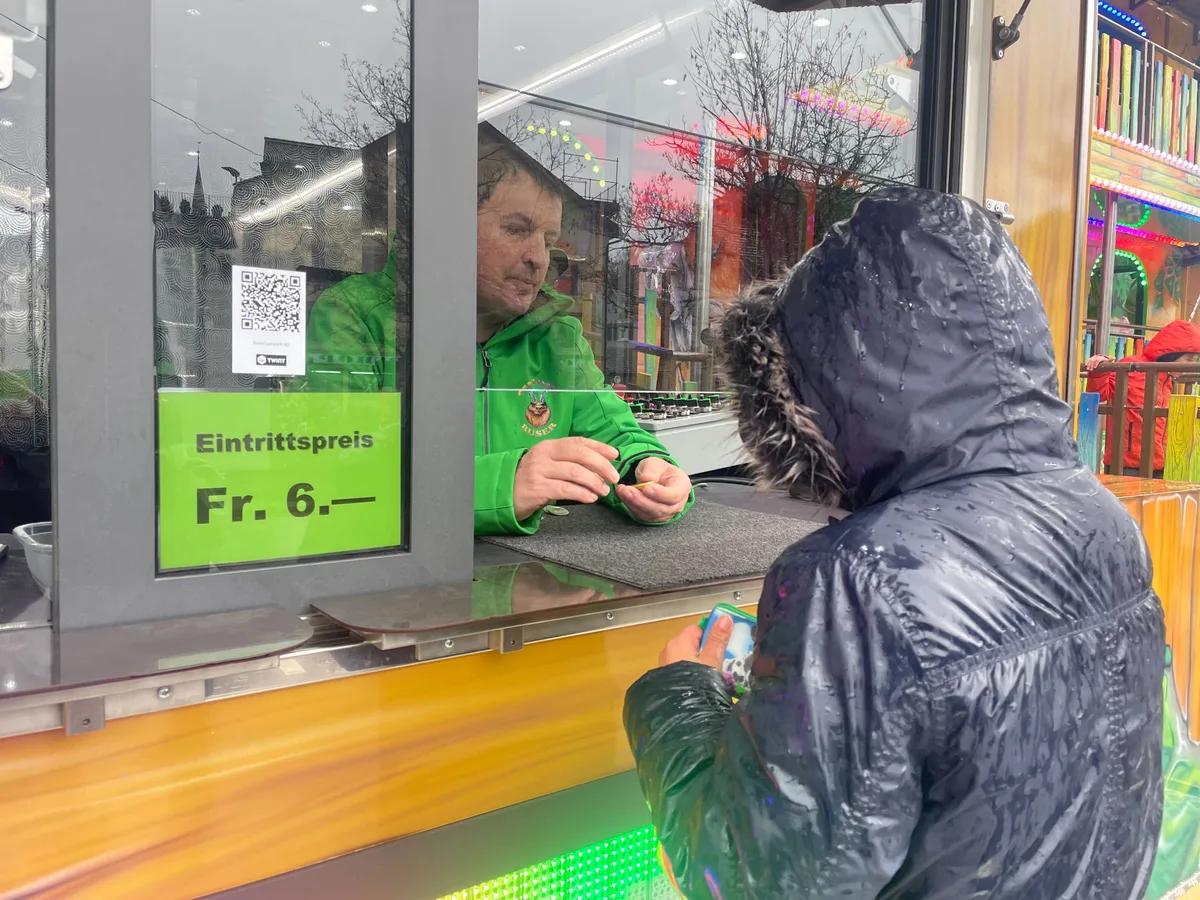 Kind (von hinten) steht an einem Kassenhäuschen und kauft bei einem Mann Jetons. 