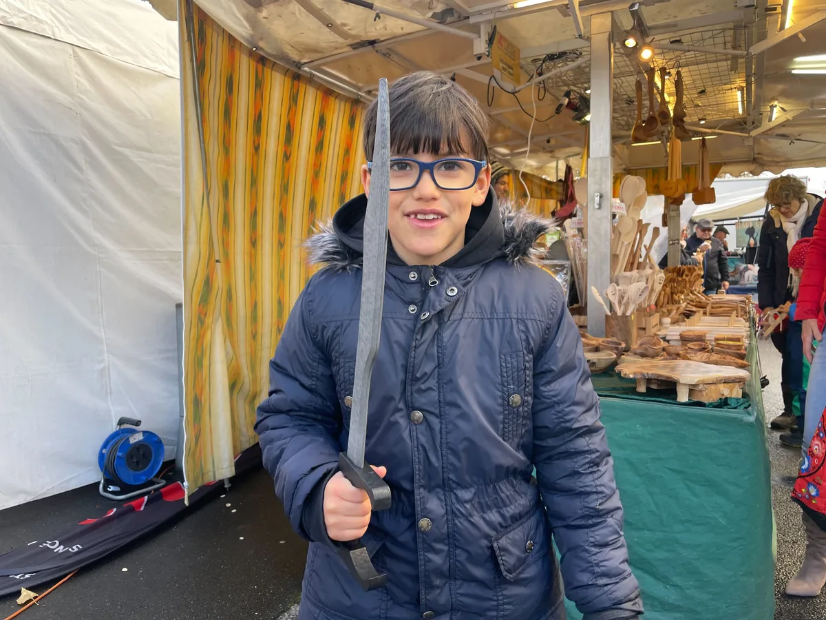 Junge mit Brille hält ein Holzsäbel in der Hand und lacht in die Kamera. 