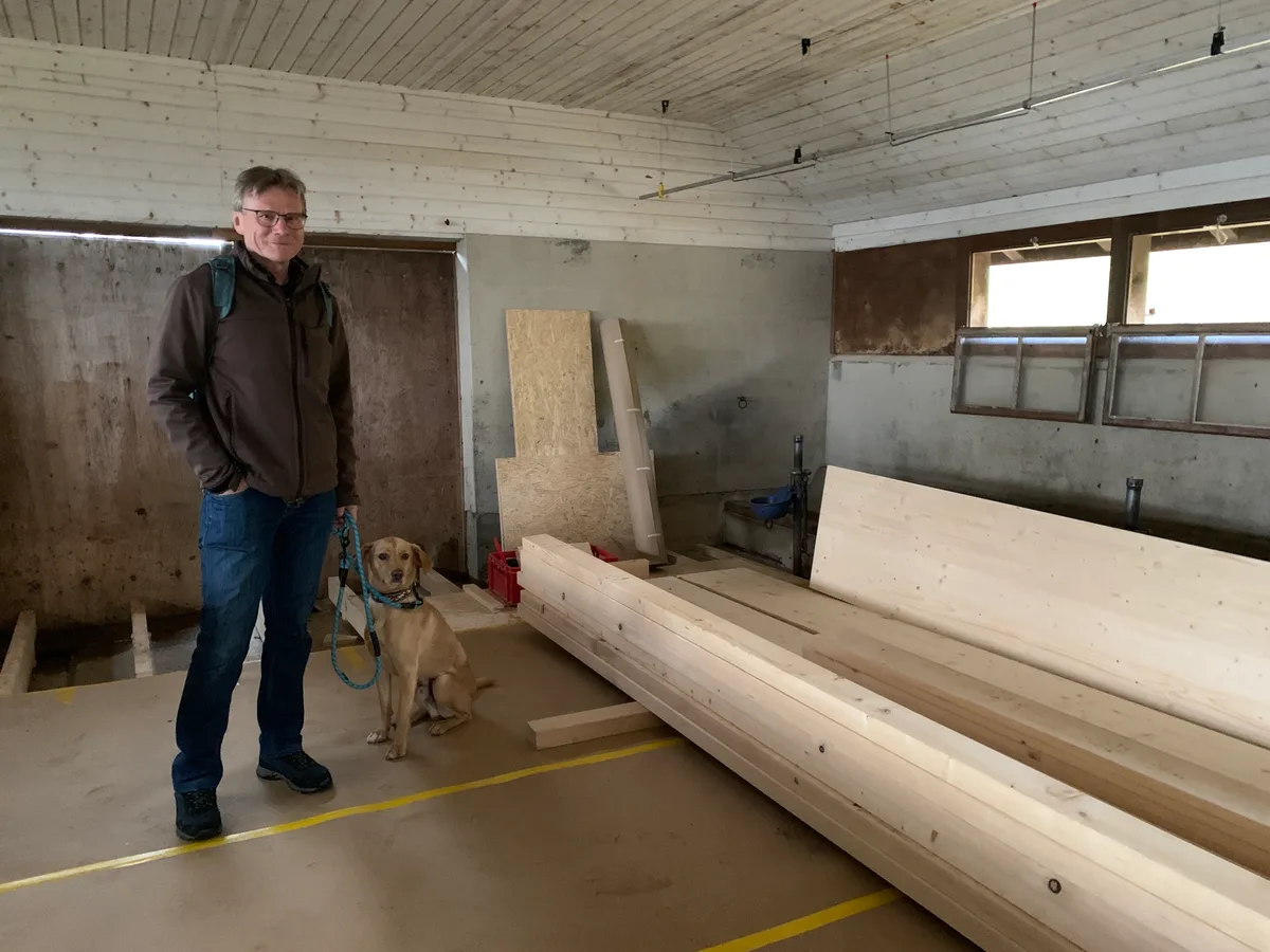 Ein Mann mit Hund steht in einem Stall neben einem Stapel Holz. 