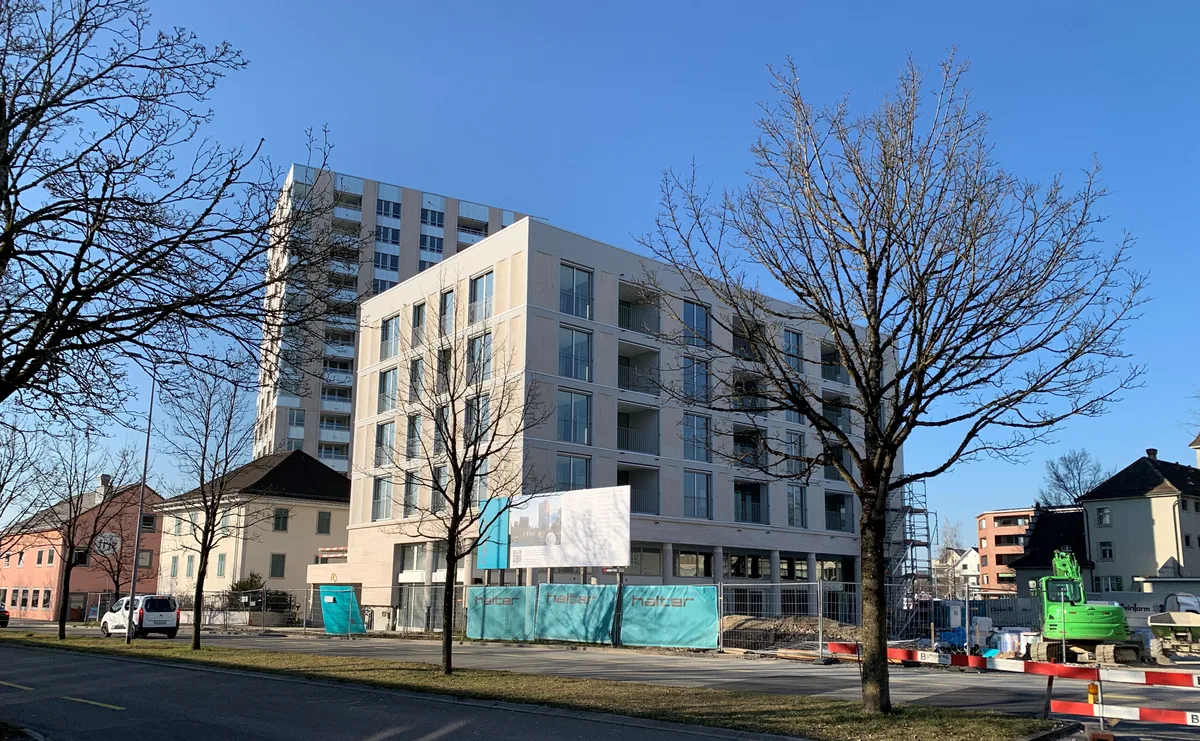 Nun werden die Umgebungsarbeiten bei der Grossüberbauung Gerichtsplatz – hier von der Zürichstrasse aus gesehen – in Angriff genommen. Grosse Wohnblöcke umschliessen zwei kleinere Häuser und daneben steht ein Bagger.