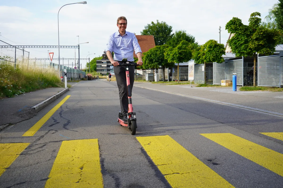 Mann im Hemd fährt mit Scooter an einen Fussgängerstreifen.