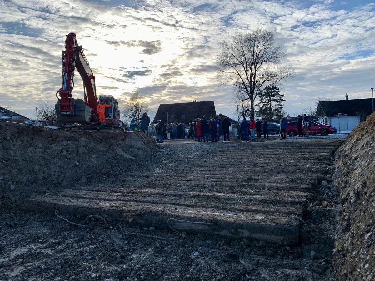 Blick aus einer Baugrube auf einen Bagger und eine Gruppe von Leuten.