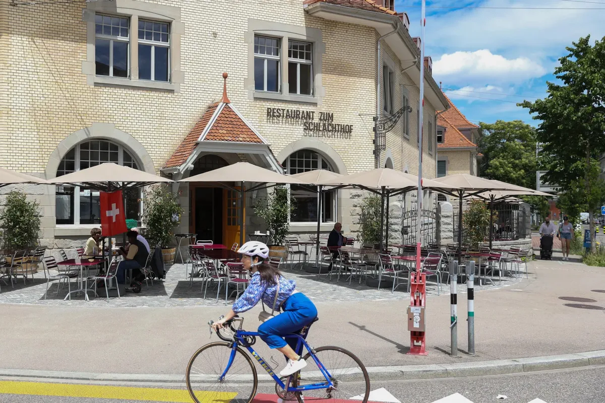 Traditionsrestaurant beim Letzigrund: Das Restaurant zum Schlachthof soll für die Hinwiler Gastrounternehmer das Tor zur Stadt Zürich sein. (Archiv) Aussenansicht des Restaurants zum Schlachthof in Zürich. Man sieht eine helle Backsteinfassade und davor Tische und Stühle unter grossen Sonnenschirmen. Eine Radfahrerin fährt durchs Bild.