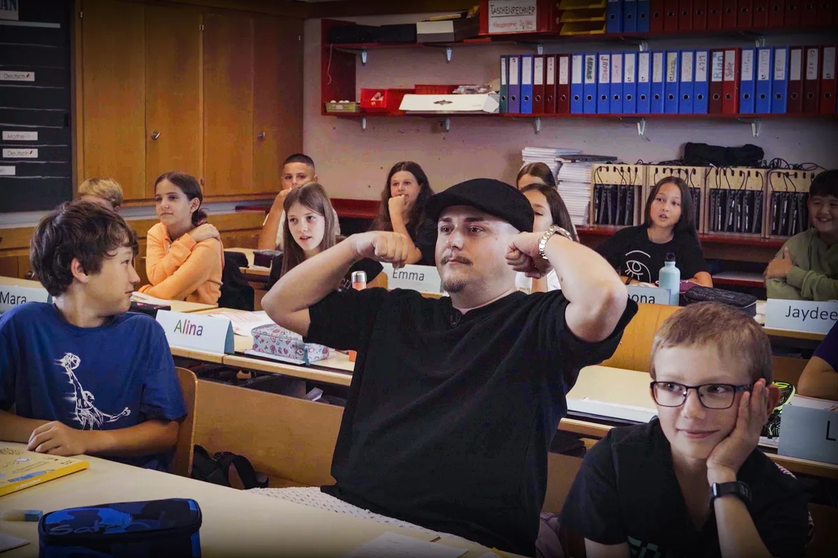 Hinter Schulbänken sitzen Kinder und ein Mann.