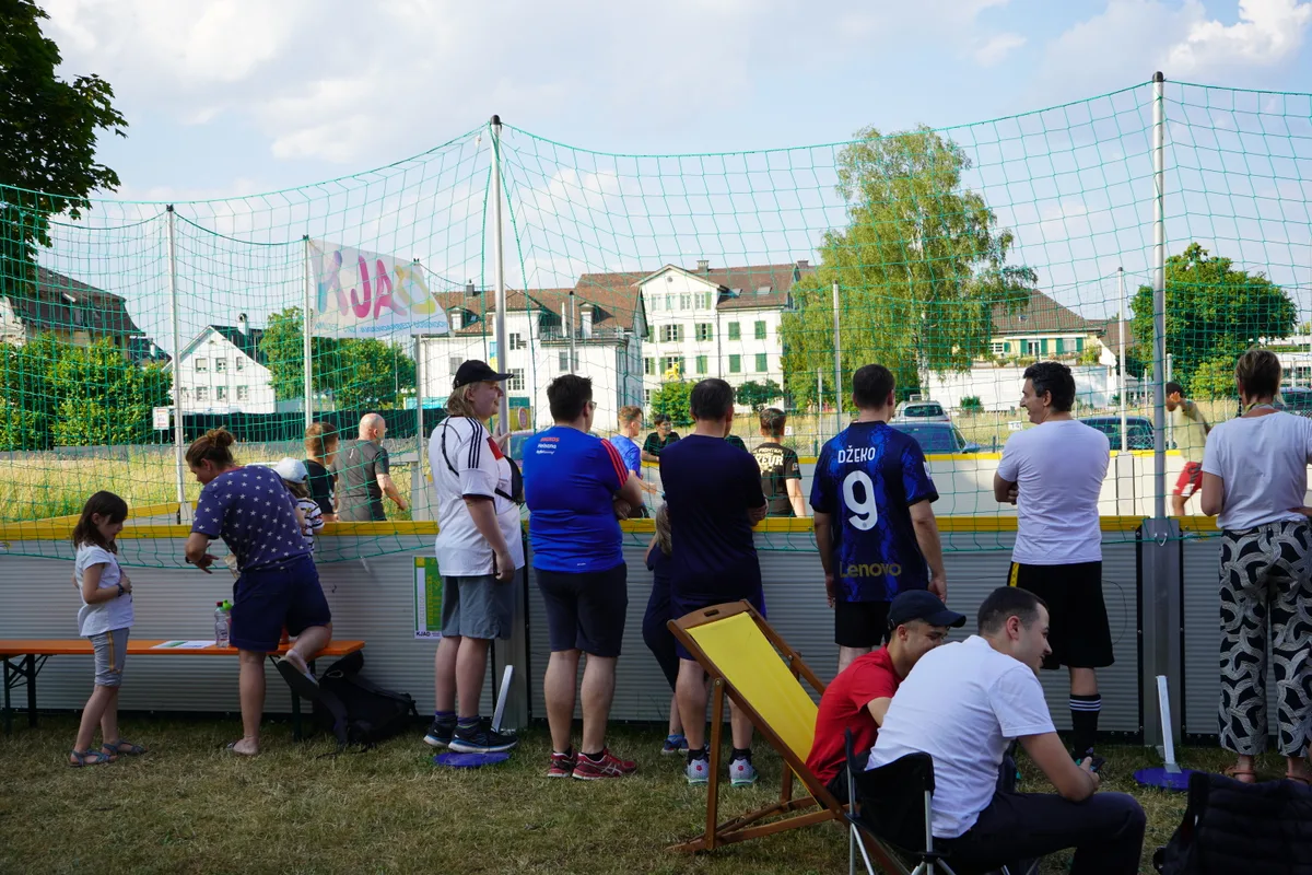 Ein Haufen Menschen neben einem umzäunten Fussballfeld.