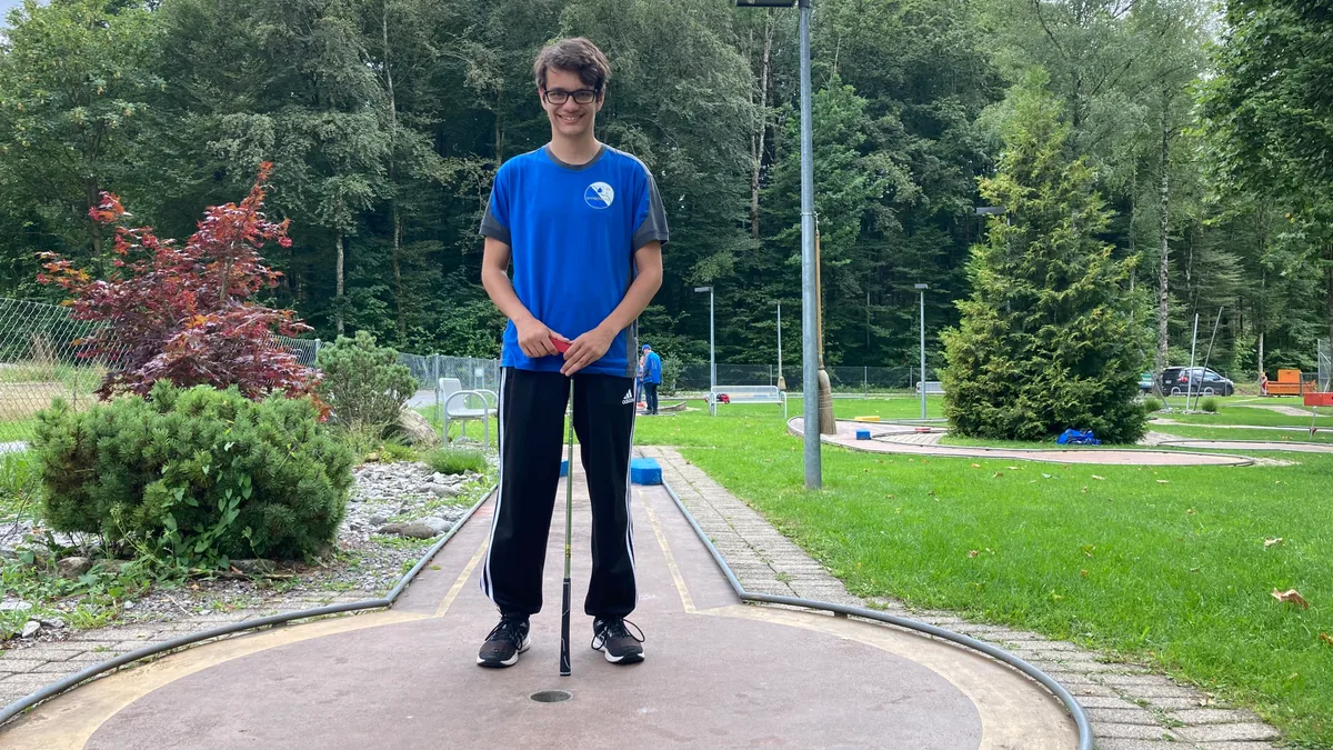 Ein junger Minigolfspieler steht lächelnd auf einer Bahn.