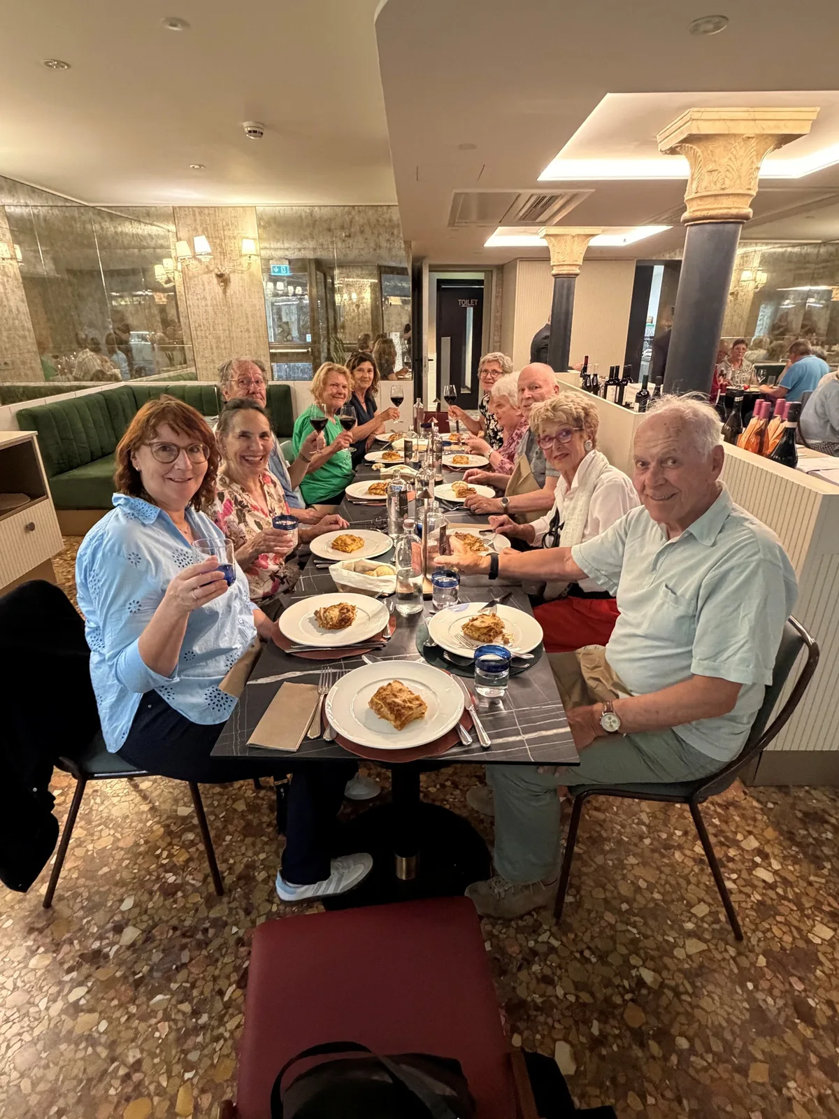 Erstes gemeinsames Abendessen in einem Restaurant nahe beim Hotel. Menschen sitzen an einem langen Tisch, Teller mit Lasagne vor sich und Gläser in der Hand.