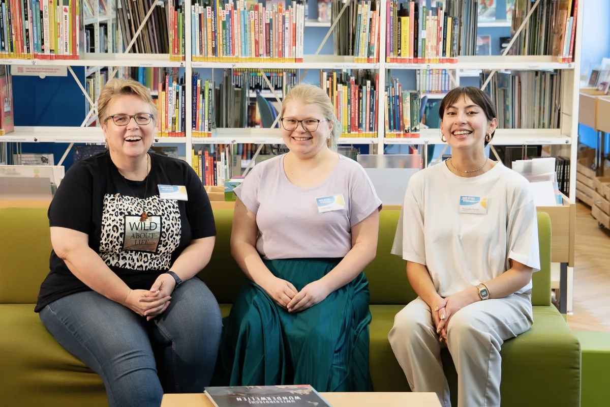 Kommt während der Arbeit eigentlich nicht zum Sitzen: Das Bibliotheksteam um Bettina Asch (links). Drei Frauen sitzen auf einem Sofa in einer Bibliothek und lächeln in die Kamera.