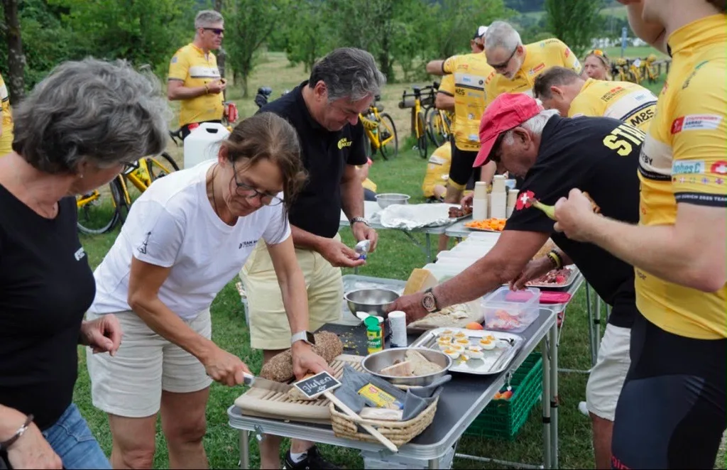 Die Helfenden sind dafür besorgt, dass die Velofahrer unterwegs Energie tanken können. Man sieht Helfer und Velofahrer um ein Snackbuffet stehen.