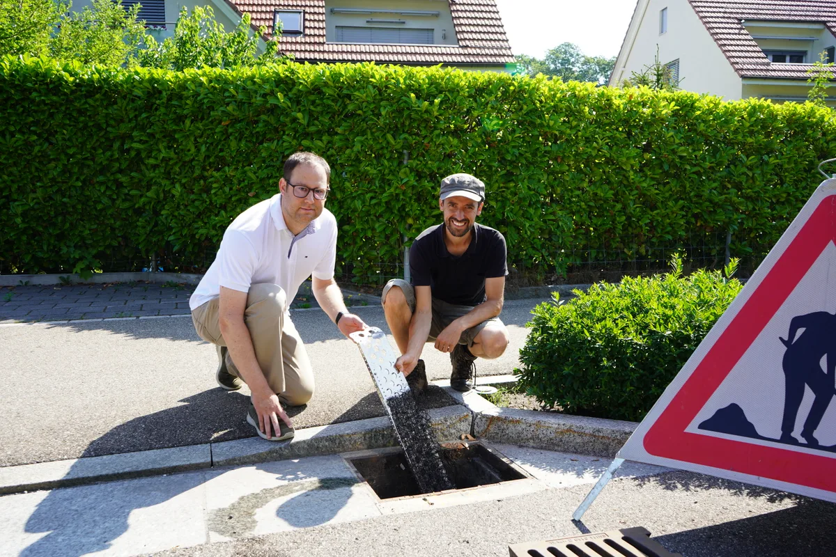 Zwei Männer knien vor einem offenen Schacht und halten ein Stück Metall in den Händen.
