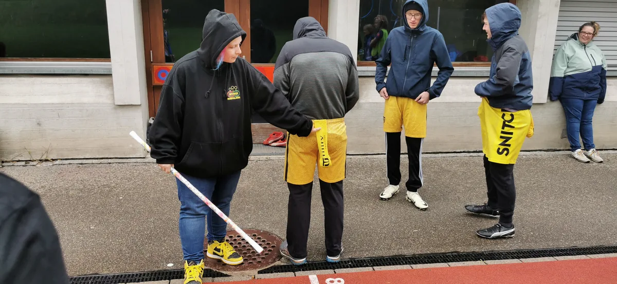 Der Flag steckt in einer Stoffsocke, die mit Klettverschluss an einer Hose befestigt ist. Linnéa Grimm erklärt der Mannschaft eine gute Taktik, den Flag während des Spiels schnell zu packen. Linnéa Grimm zeigt auf den Flag an einem Spieler.
