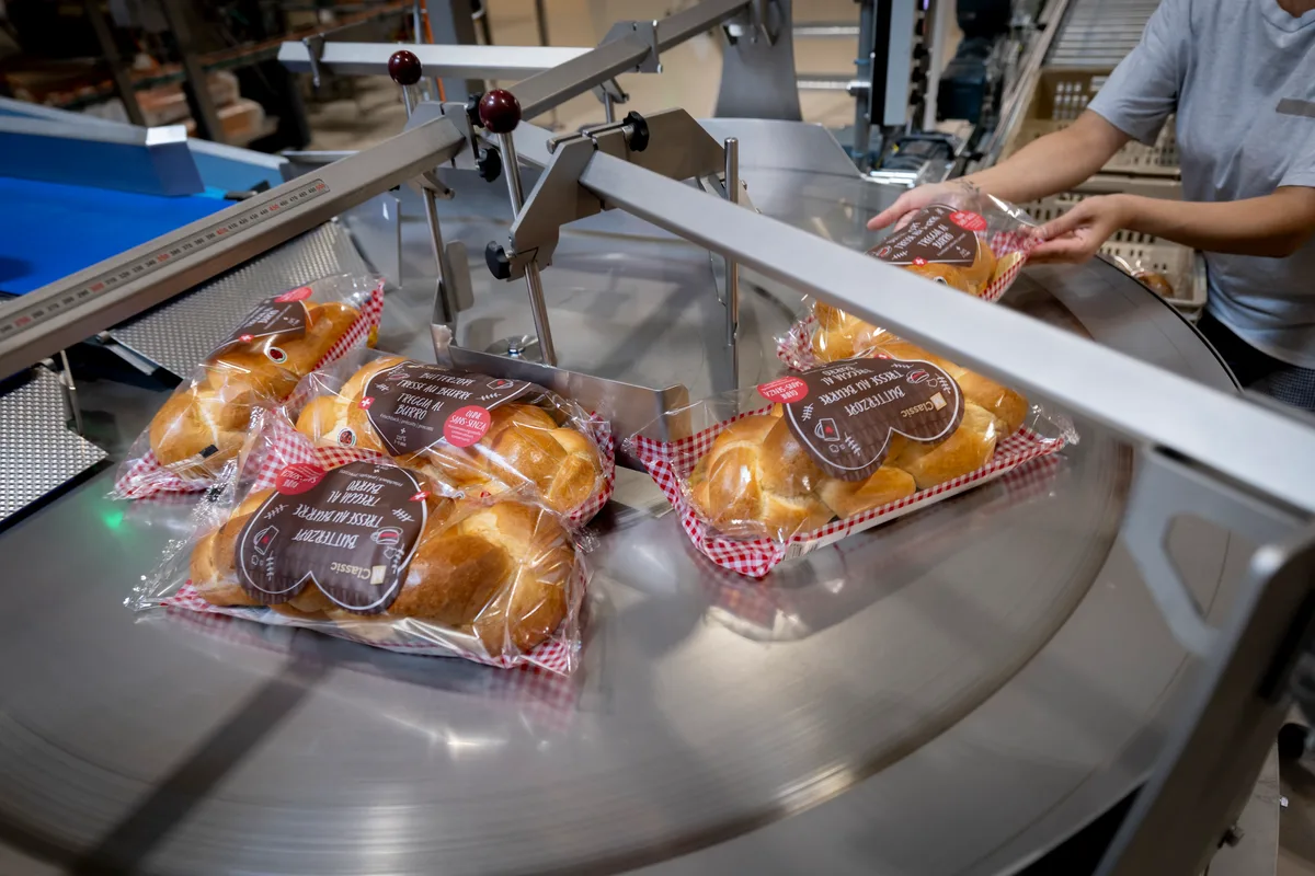 Die gebackenen Zöpfe werden nach dem Abkühlen maschinell eingepackt. Eine Arbeiterin greift sie dann vom Band, um sie in Kisten zu verfrachten. Eine Arbeiterin greift Zöpfe vom Band, um sie in Kisten zu verfrachten.
