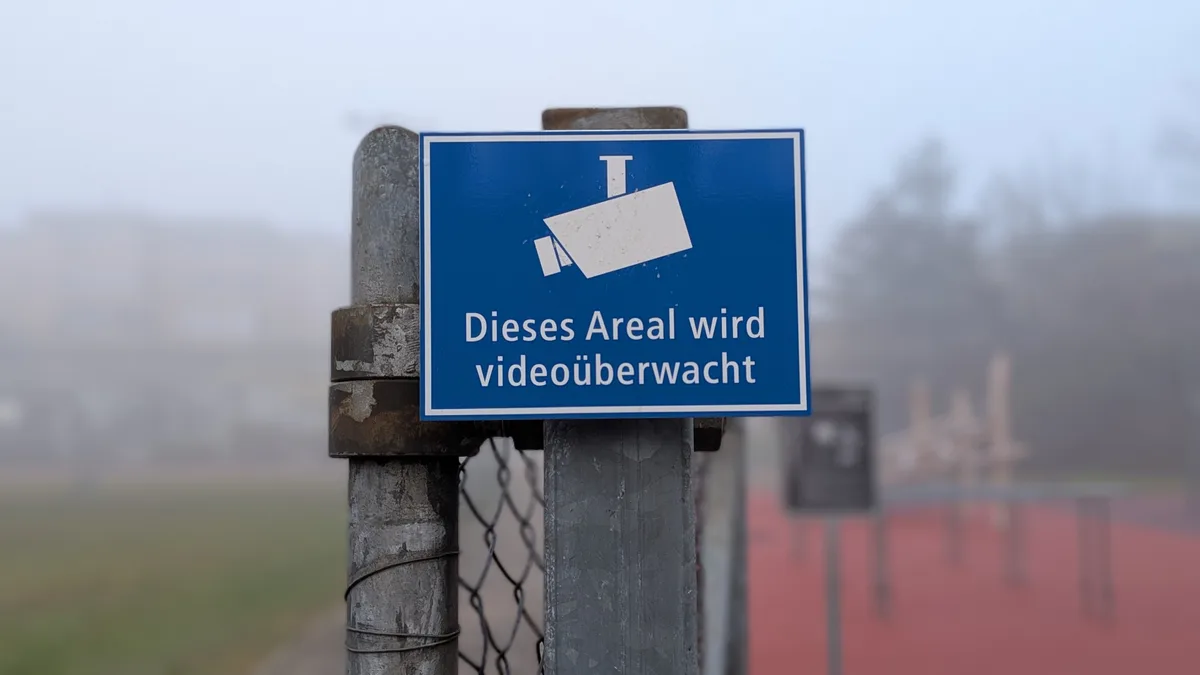 Man sieht ein blaues Schild mit der Aufschrift «Dieses Areal wird videoüberwacht», im Hintergrund im Nebel eine Schulanlage.