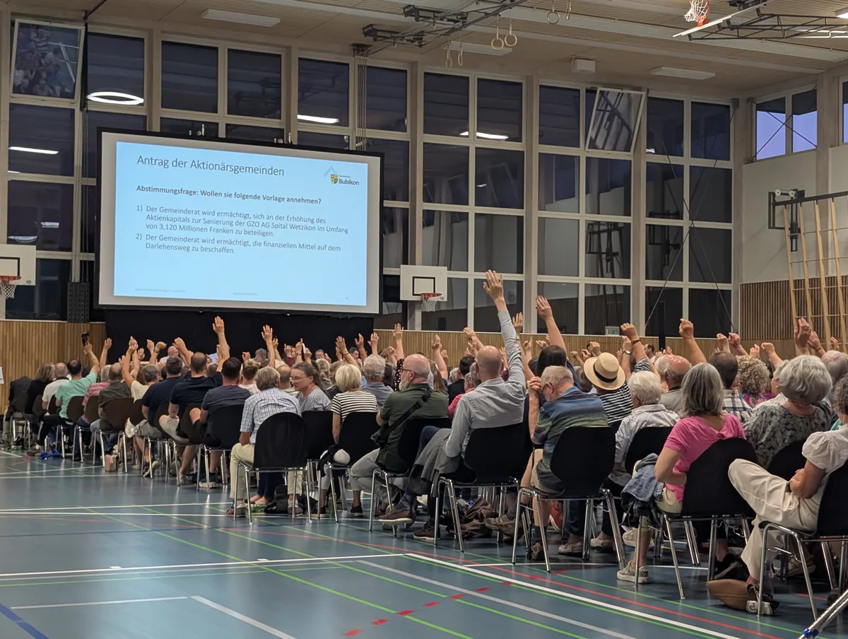 Man sieht zahlreiche Leute, die in einer Turnhalle sitzen und den Arm in die Höhe strecken.