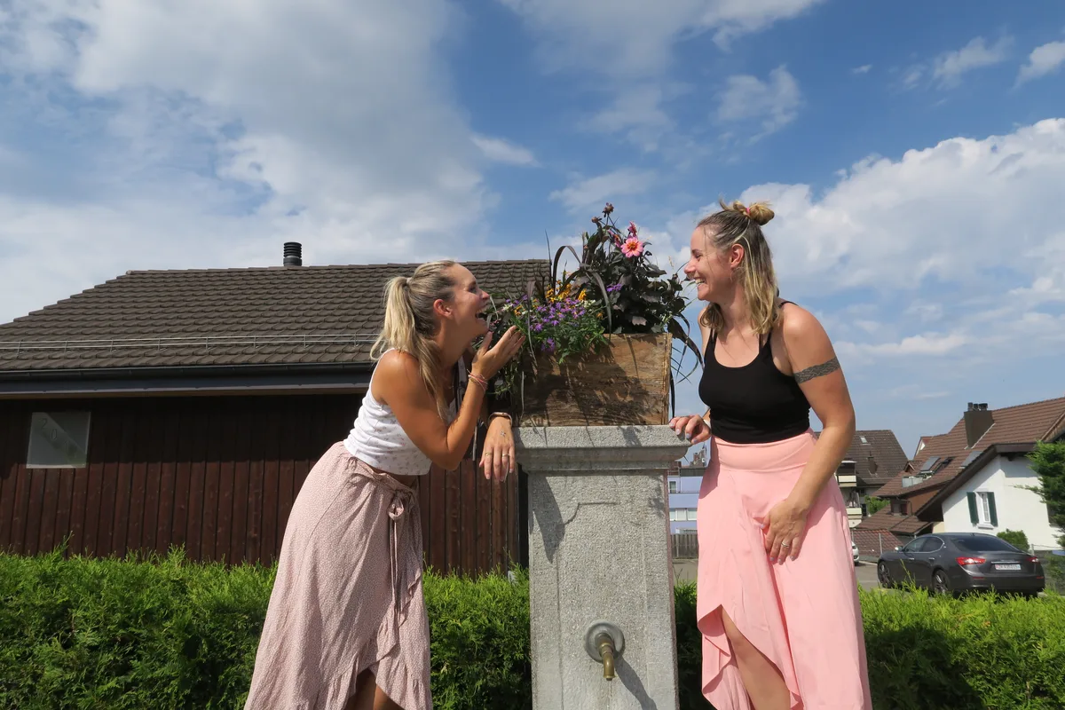 Das Bild zeigt Vanessa Ender und Ramona Bucher, die auf dem Brunnenrand stehen und an den Blumen riechen.