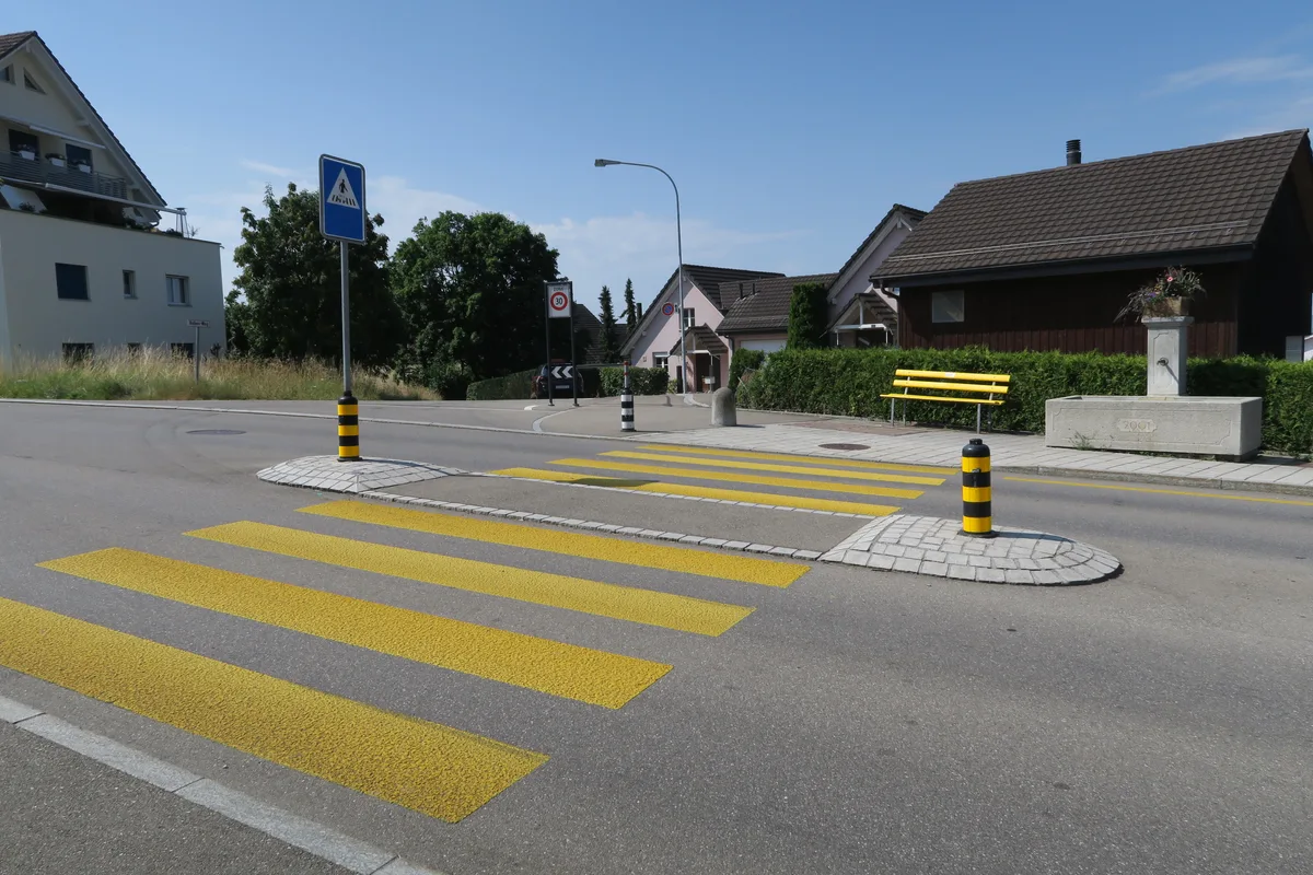 Das Bild zeigt eine Strasse und dahinter ein gelbes Bänkli und einen Brunnen.