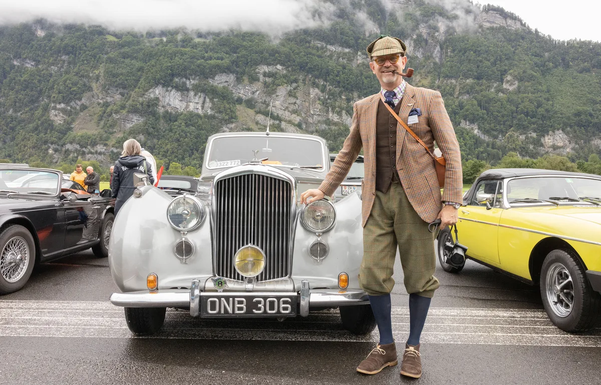 Ein Mann steht vor seinem Rolls-Royce-Oldtimer. Er trägt Mode aus der ersten Hälfte des letzten Jahrhunderts, unter anderem karierte Knickerbocker.