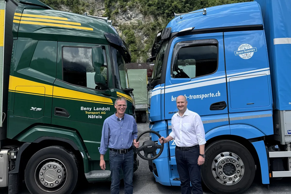 Zwei Männer stehen vor einem grünen und einem blauen Lkw und halten gemeinsam ein Lenkrad.