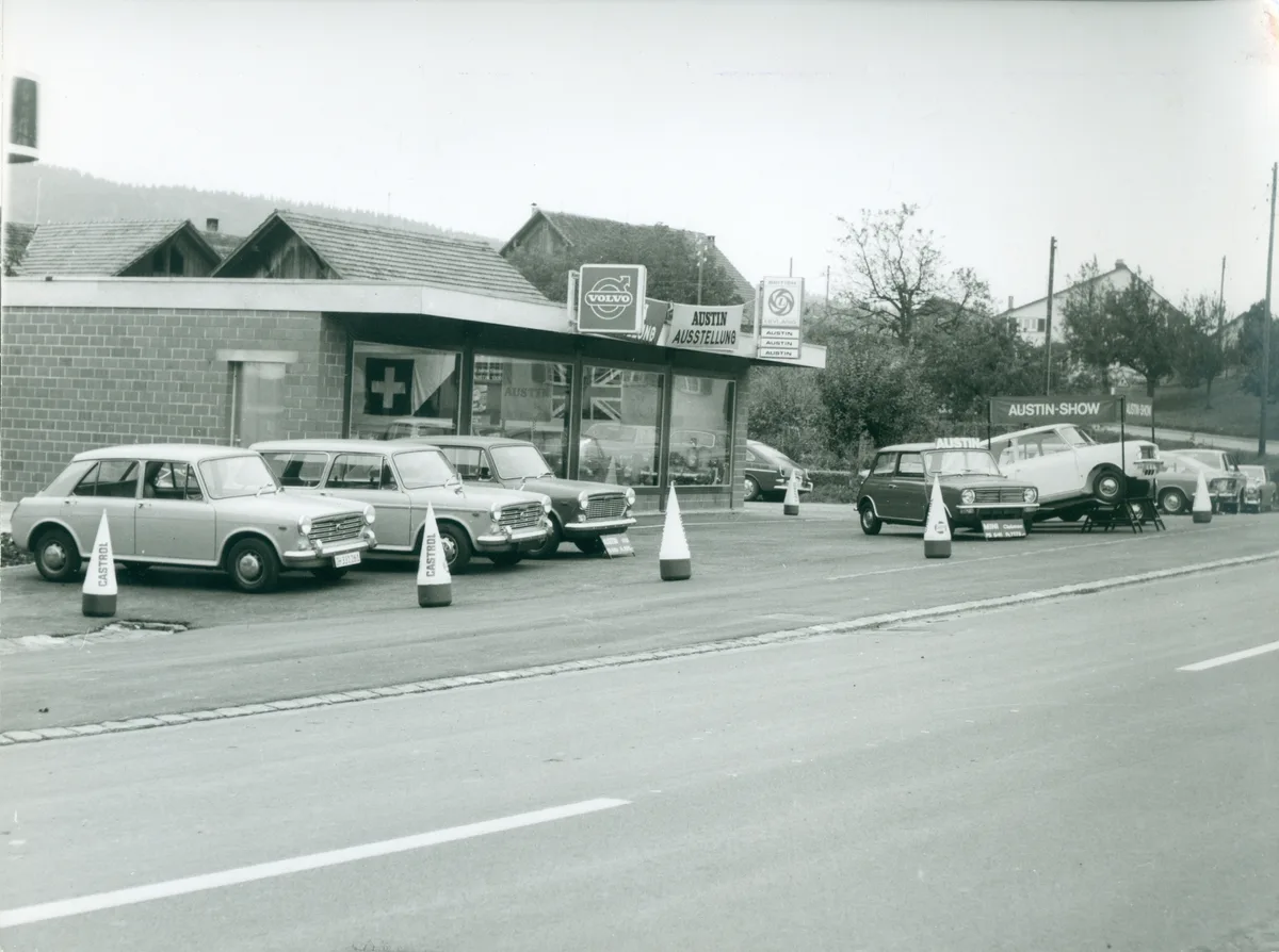 Schwarz-Weiss-Bild der Garage Glättli in Hittnau aus dem Jahr 1970. Man sieht ein Werkstattgebäude, davor stehen Neuwagen der Marke Austin.