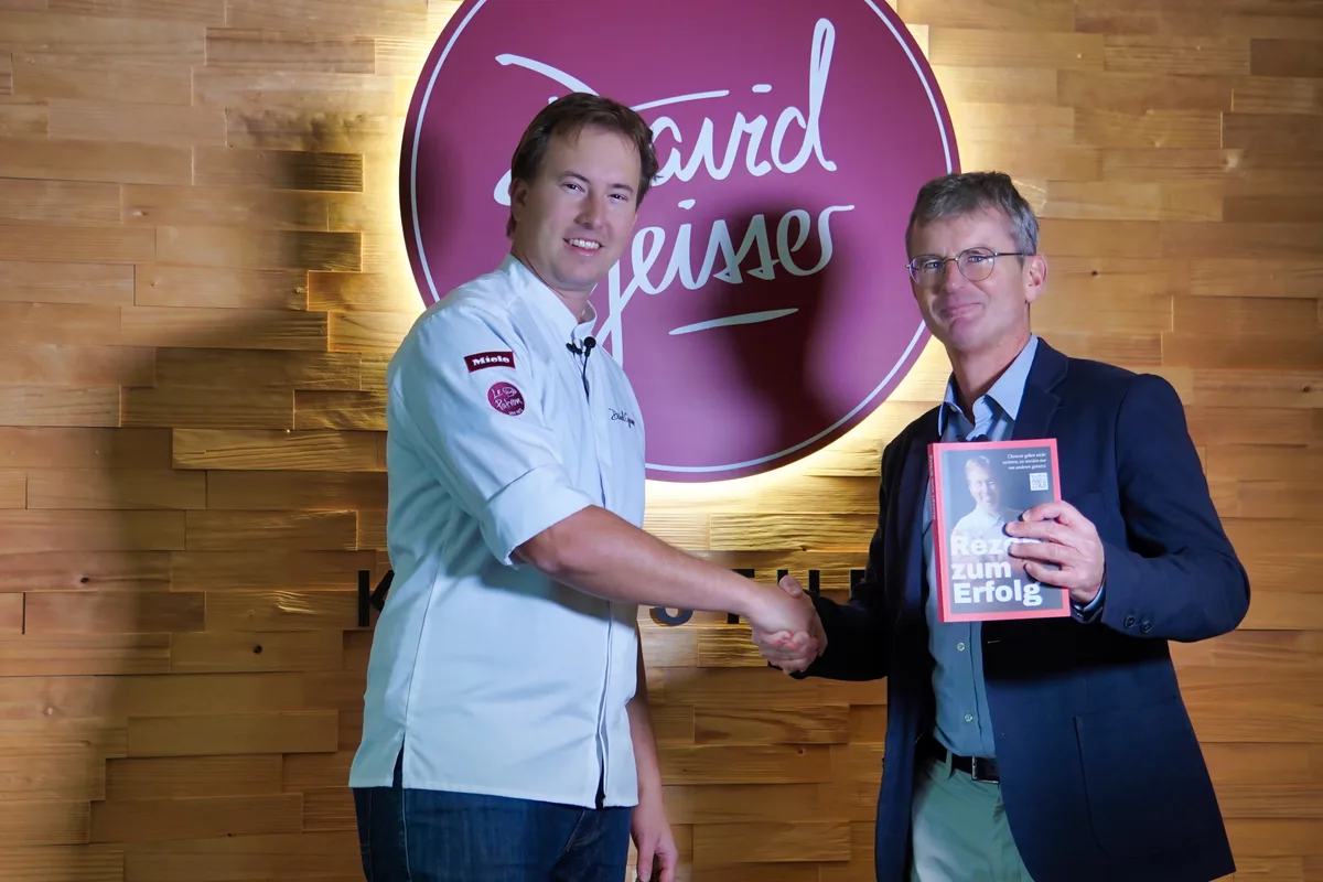 Koch David Geisser und Autor Thomas Renggli schütteln die Hände, Renggli hält ein Buch in der Hand.